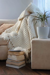 A comfortable living room featuring a beige linen sofa draped with a thick, cream-colored wool blanket. A textured jute rug covers the floor, and a reclaimed wood side table holds a potted plant.