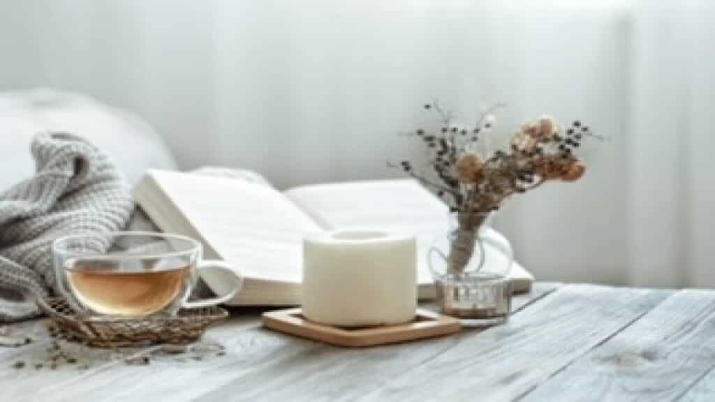 Glass teacup next to a comfy cozy home candle with an open book